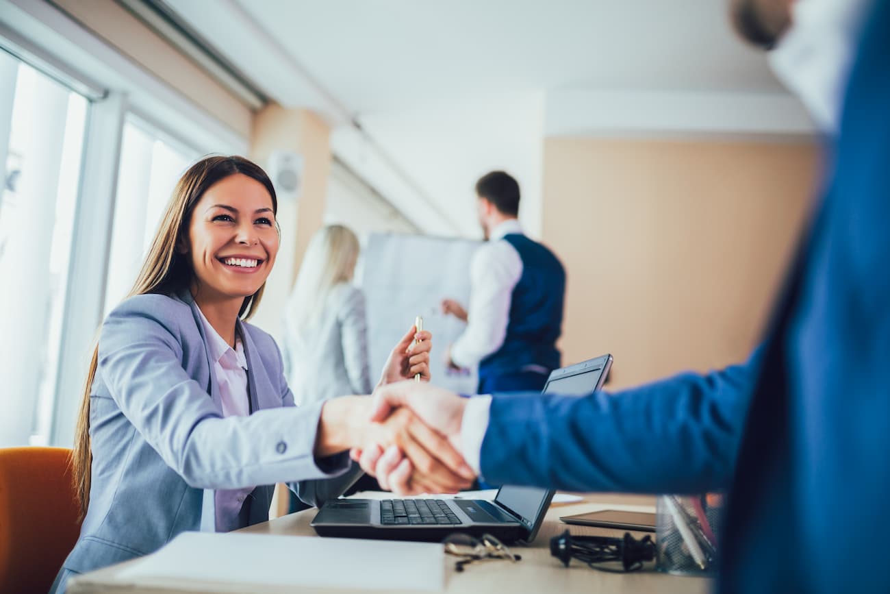 Lächelnde Geschäftsfrau schüttelt die Hand eines Geschäftspartners im Büro, während Kollegen im Hintergrund arbeiten, symbolisiert erfolgreichen Abschluss und Teamarbeit.