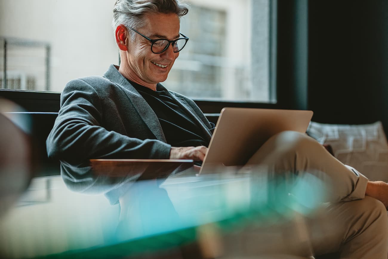 Lächelnder Mann sitzt entspannt auf einem Sofa und arbeitet an seinem Laptop, symbolisiert die Notwendigkeit der Angabe persönlicher Profildaten zur Identifikation und Einladung zu Projekten.