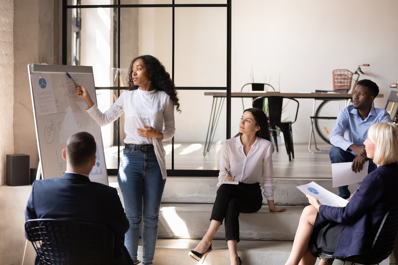 Frau präsentiert Pläne auf einem Whiteboard vor einem Team in einem modernen Büro, symbolisiert sorgfältige Planung und Zusammenarbeit für Crowdtesting-Projekte.