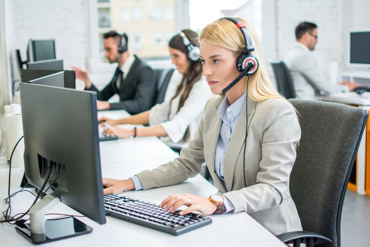 Frau mit Headset arbeitet am Computer in einem Büro, Kollegen im Hintergrund, symbolisiert sorgfältige Auswahl und Durchführung von Tests und Überwachung des Projektfortschritts.