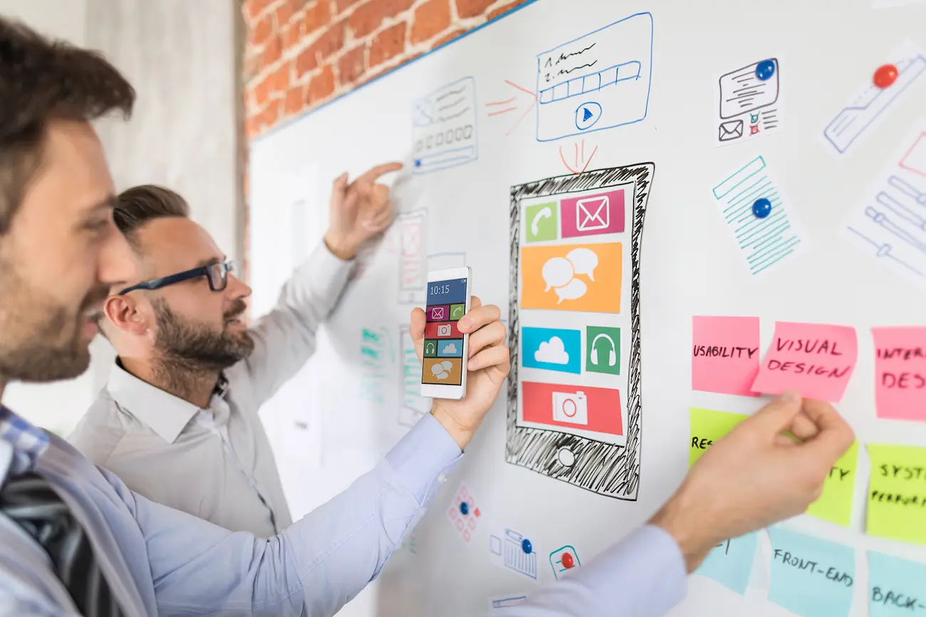 Zwei Männer planen Testabläufe auf einem Whiteboard, während einer ein Smartphone hochhält. Die Szene zeigt die Vorbereitung realistischer Testszenarien für digitale Produkte.